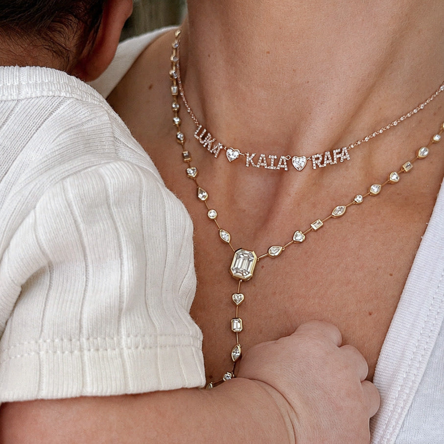 A close-up lifestyle shot of a mother and child wearing layered necklaces. The mother wears a delicate gold chain necklace with the names 'LUKA', 'KAIA', and 'RAFA' separated by small hearts, layered over a gold Y-necklace with a rectangular baguette centerpiece and heart details.