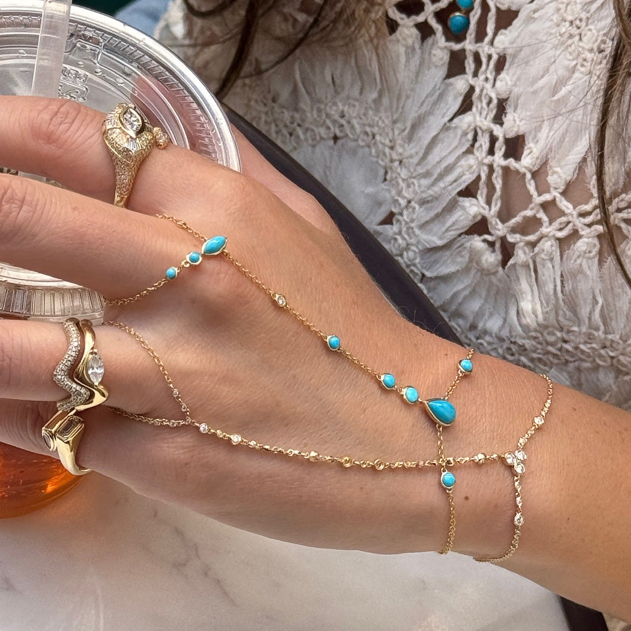 A close-up of a person's hand and wrist wearing delicate gold chain jewelry with small turquoise blue beads, including a hand chain and a bracelet, while holding a clear drink cup.