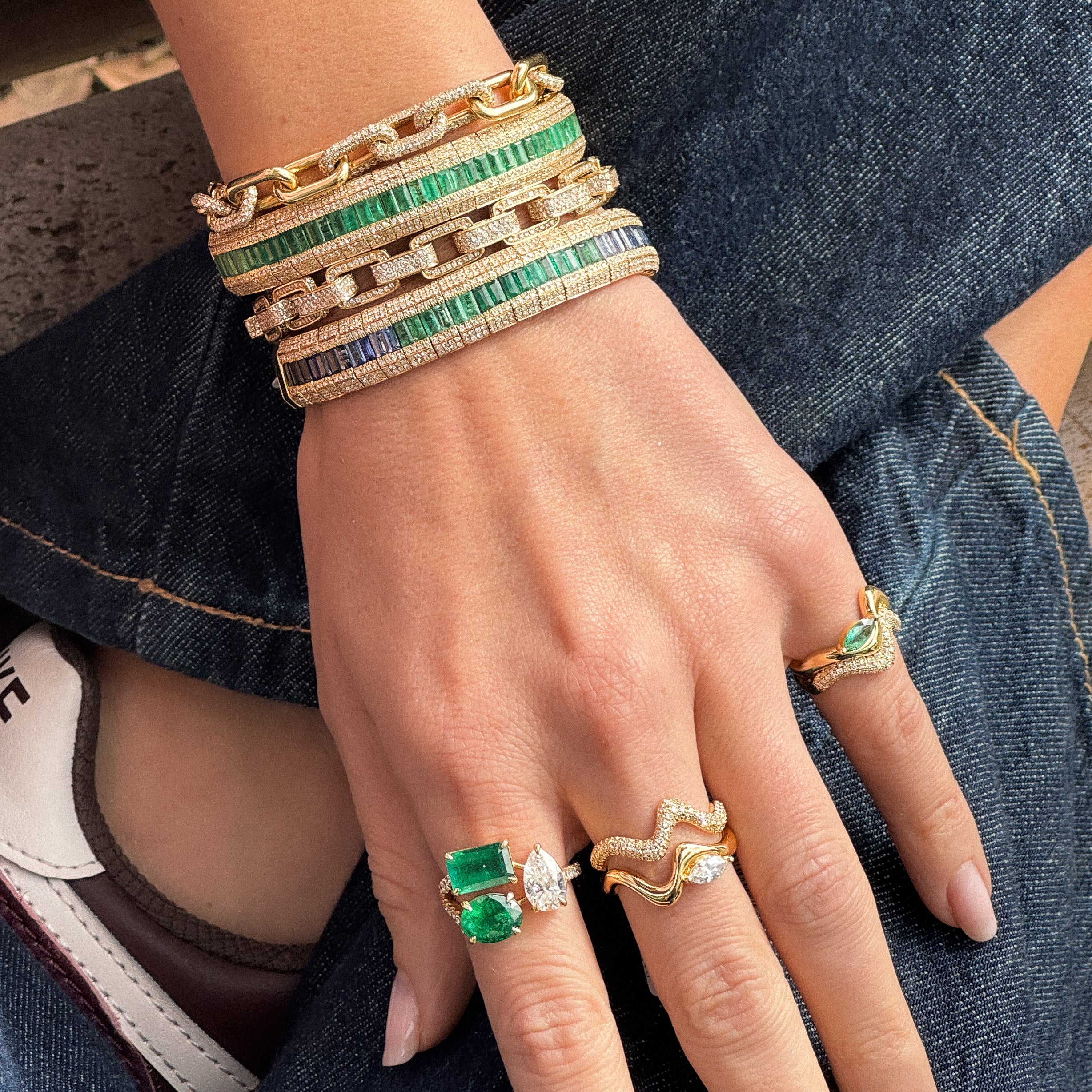 A close-up of a hand wearing multiple gold rings with green and white stones, along with stacked gold and green bracelets, set against dark denim clothing.