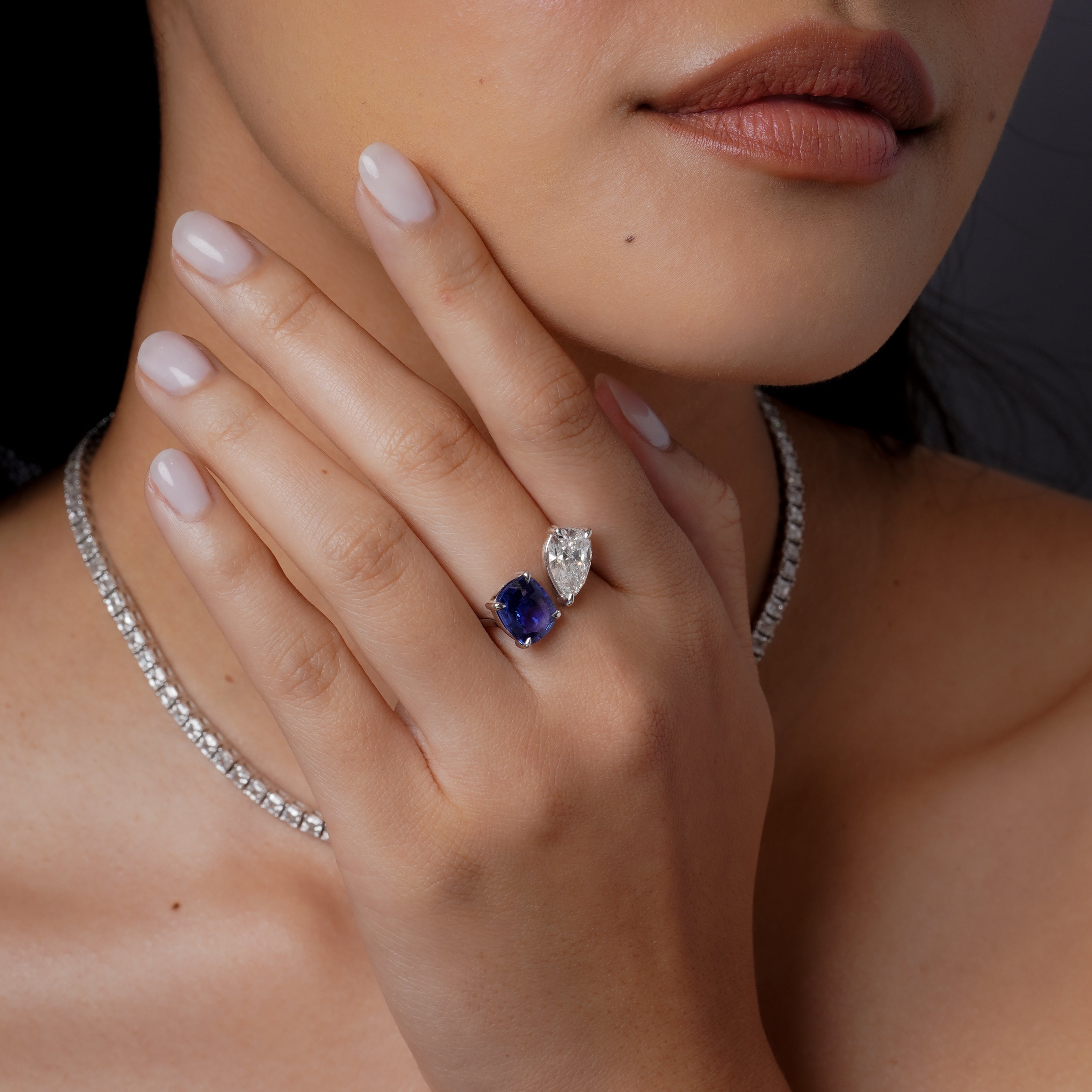 Close-up of a woman's hand wearing an 18 karat gold ring with a blue sapphire and diamond, with a necklace around her neck.