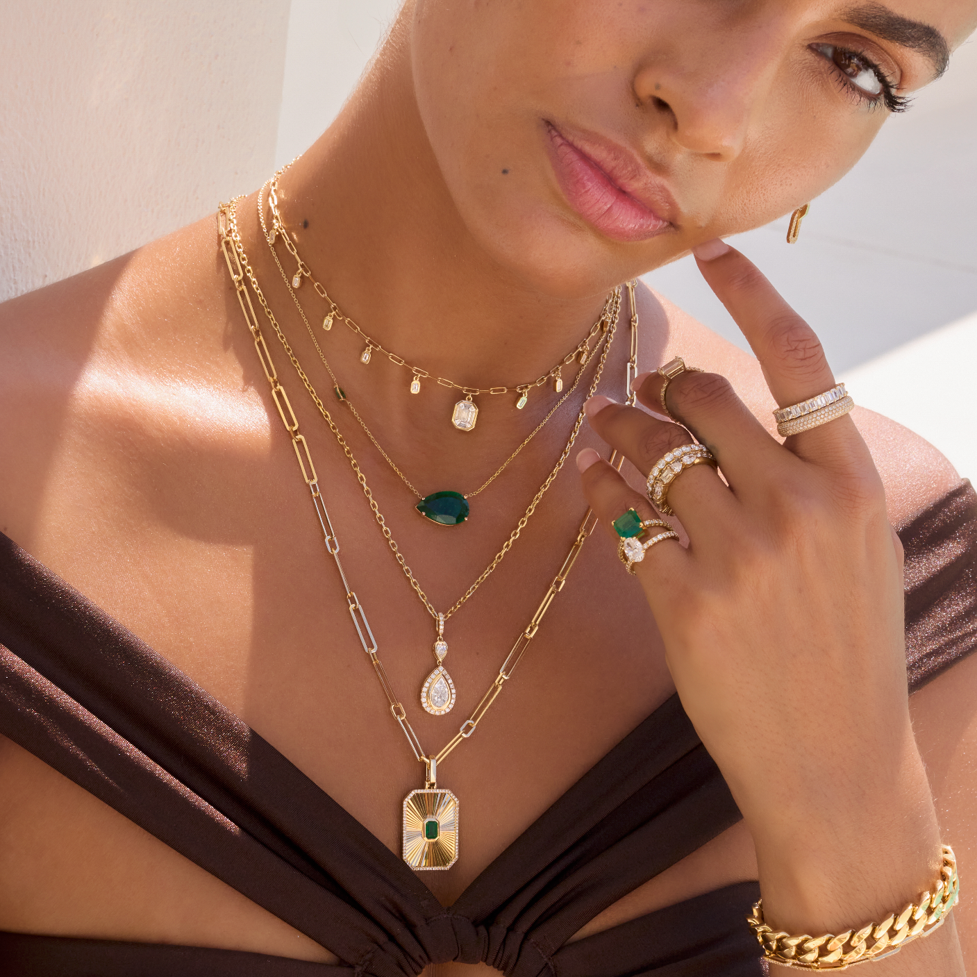 A woman wearing layered gold necklaces, rings, and a bracelet, posing next to a swimming pool. She is wearing a dark brown off-the-shoulder bikini top.