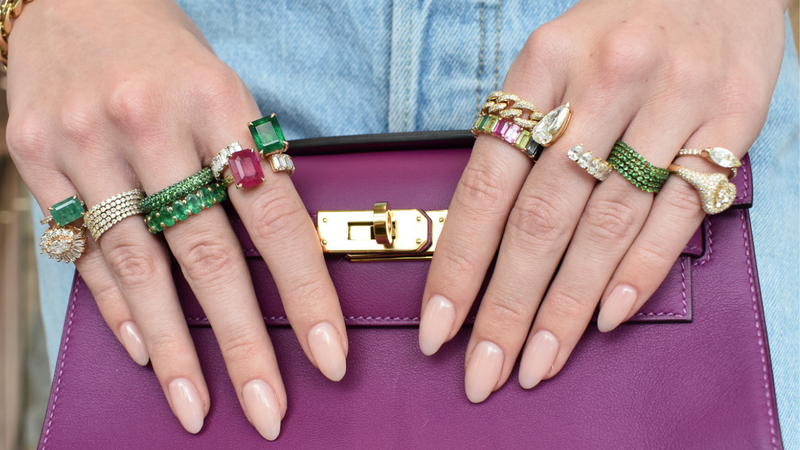 close up of hands with rings stacked with emerald and ruby gemstones holding purple clutch bag