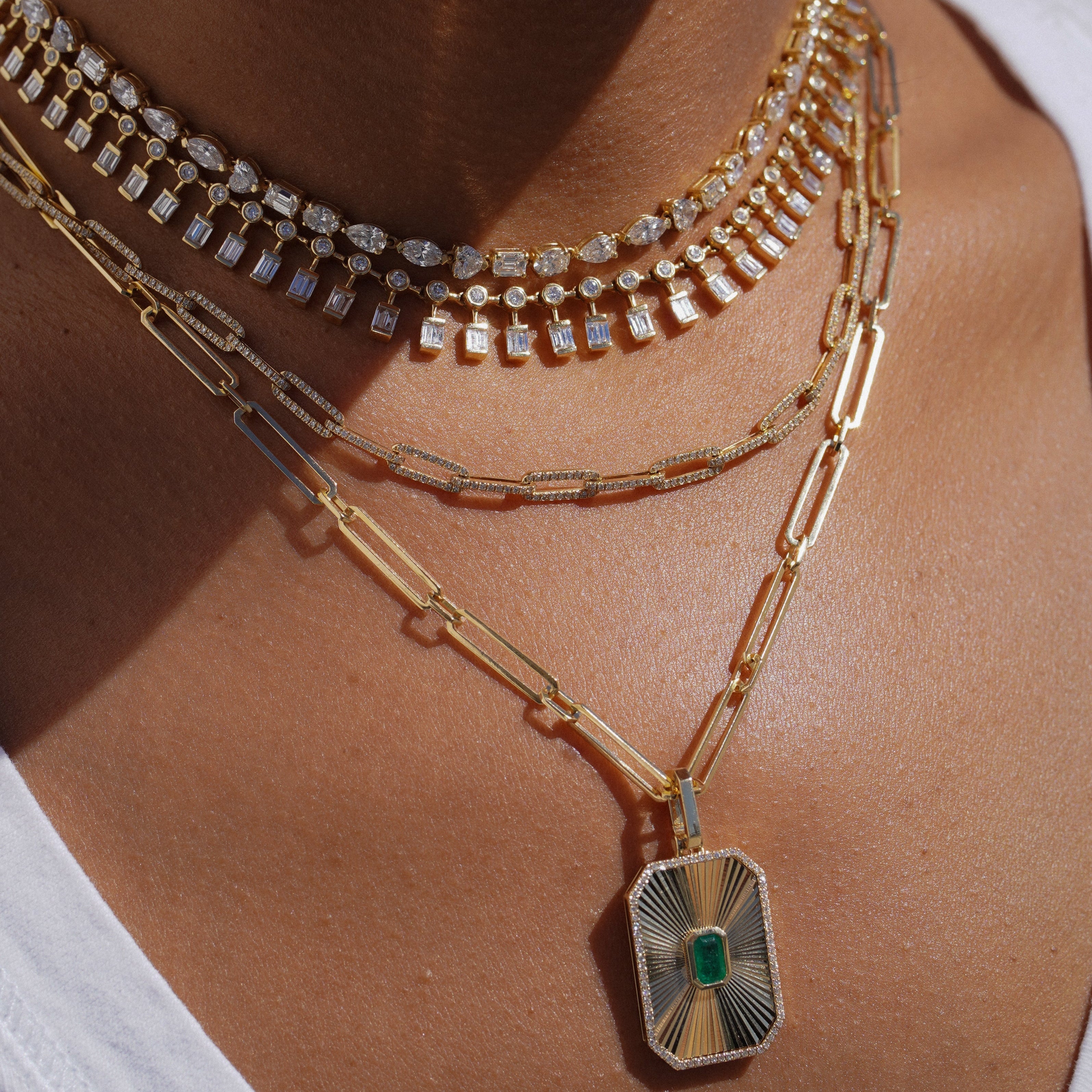 A close-up of a person's neck and chest wearing multiple layered gold necklaces, including a diamond-studded choker, a textured chain, and a paperclip chain with a rectangular pendant featuring a green center stone.