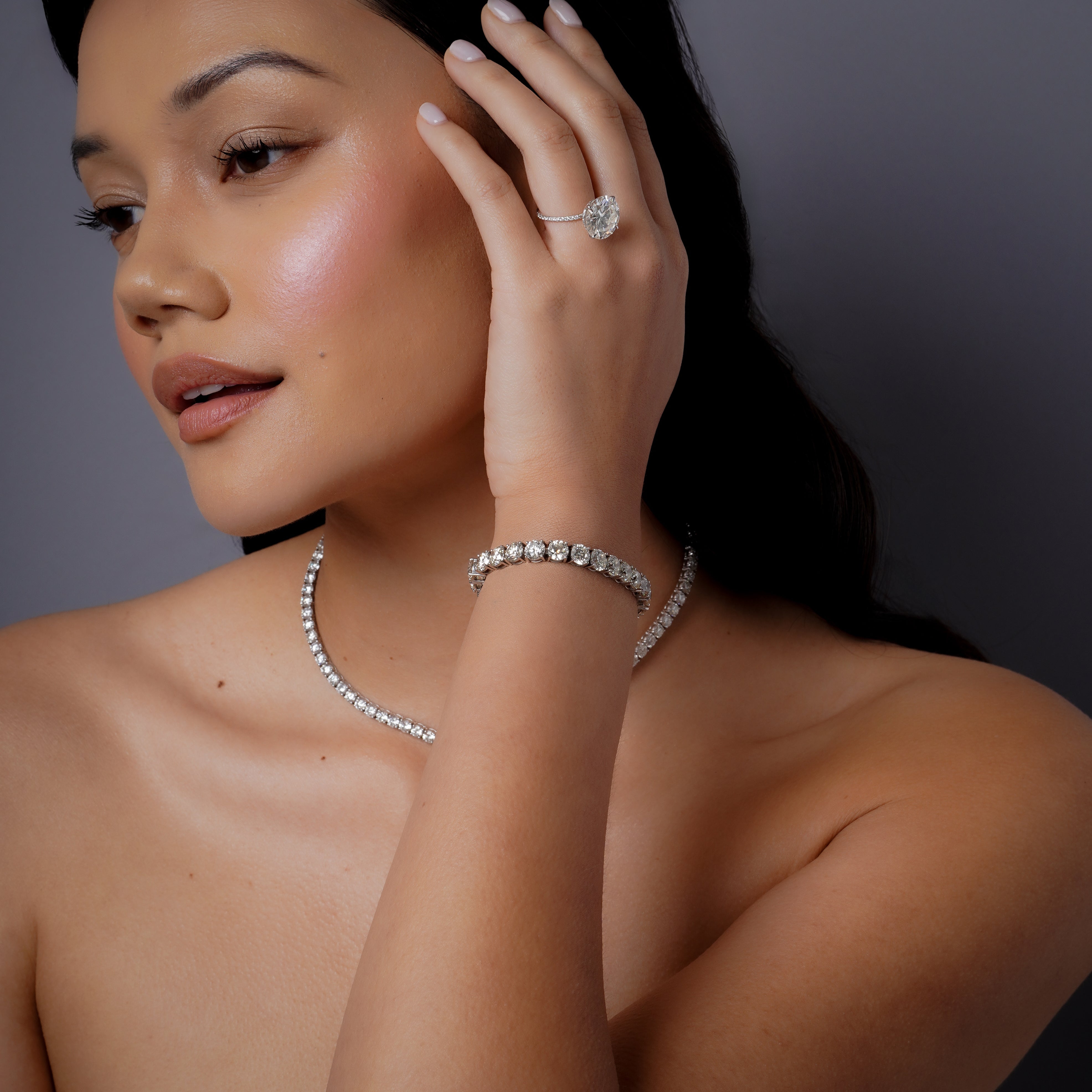 On-model lifestyle shot wearing 18k white gold diamond round tennis bracelet on wrist with matching diamond tennis necklace and diamond cocktail ring, three-quarter profile view with hand raised to face, studio lighting on grey background
