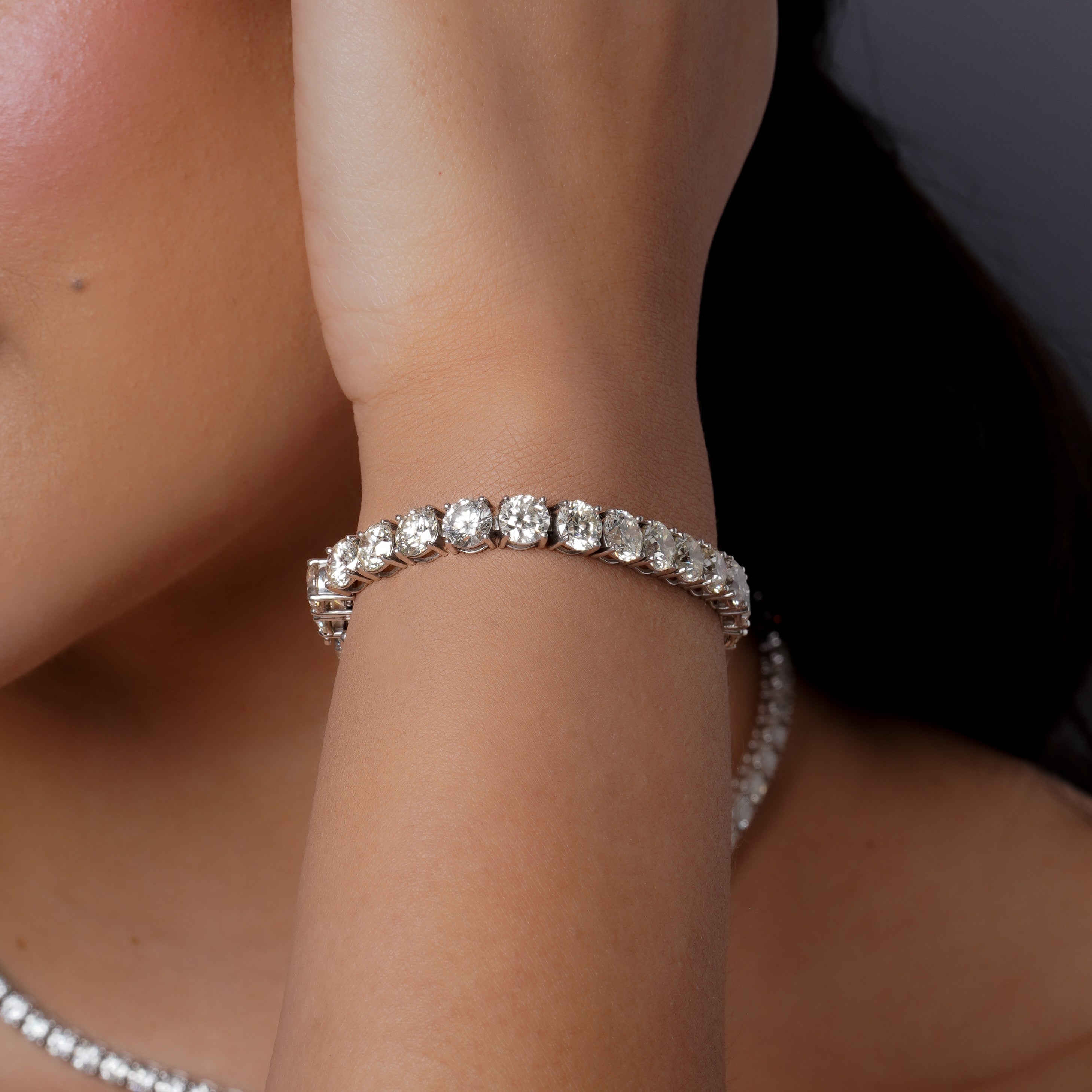 On-model wrist close-up wearing 18k white gold diamond round tennis bracelet with prong-set round brilliant diamonds in continuous line, shown at slight angle on bare wrist against dark background