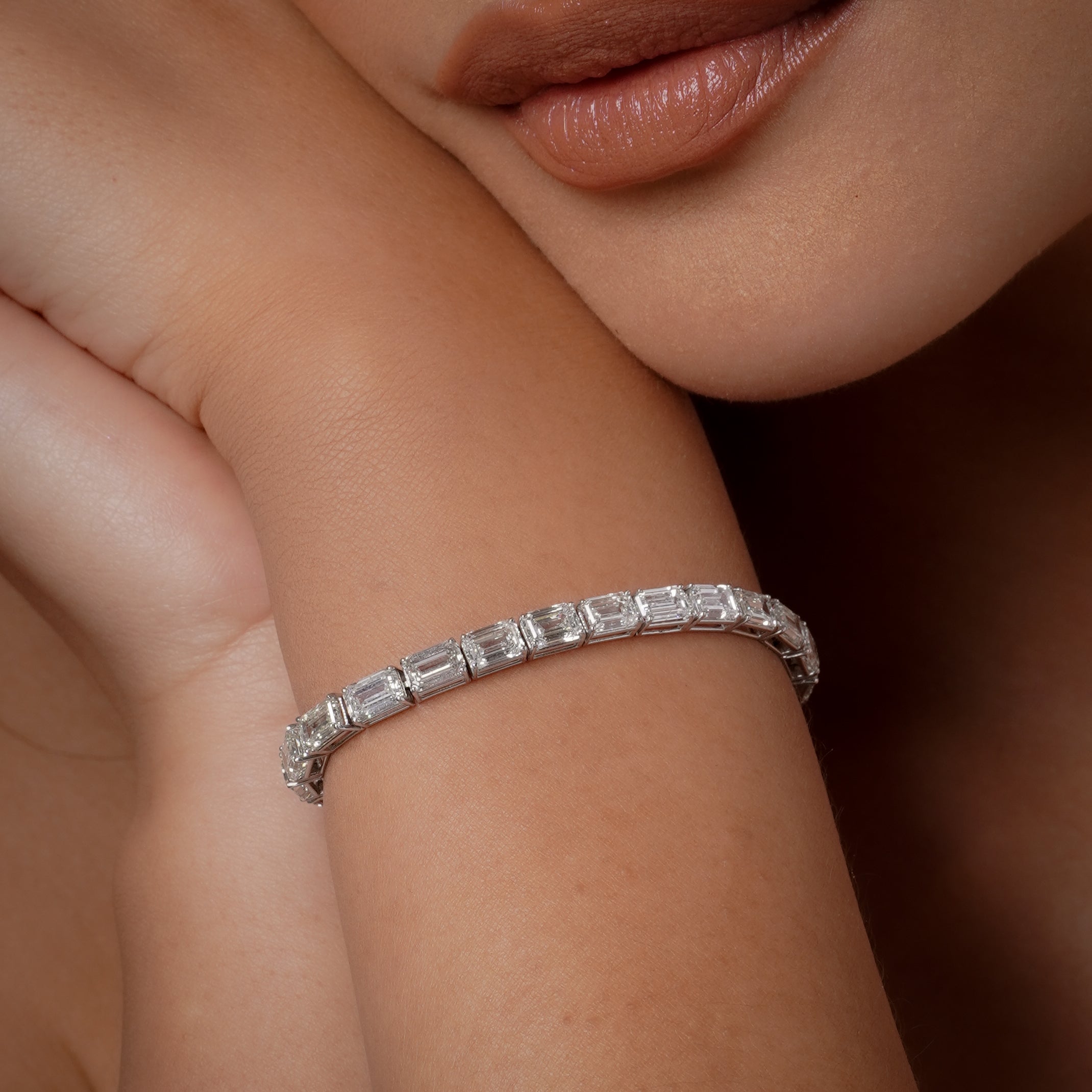 On-model wrist close-up wearing 18k white gold diamond east west tennis bracelet with alternating east-west set emerald-cut diamonds in continuous prong setting, chin and lips partially visible, studio shot on dark background