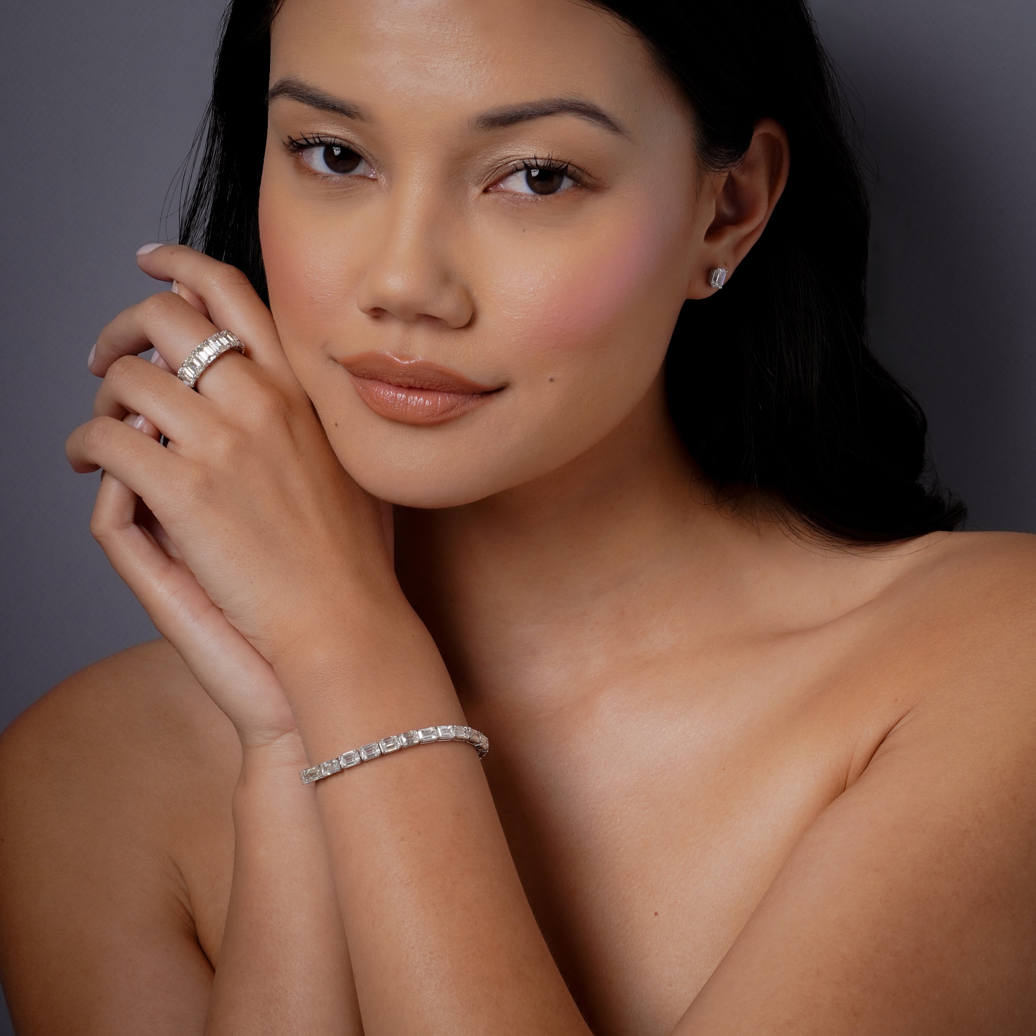 On-model lifestyle shot wearing 18k white gold diamond east west tennis bracelet on wrist with diamond stud earrings and stacked diamond rings, three-quarter profile with hands clasped near chin, studio lighting on dark background