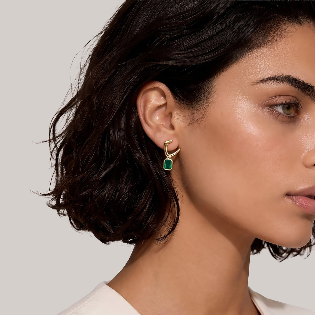 A close-up of a woman wearing a small gold hoop earring with a green rectangular stone, shown against a plain light gray background.
