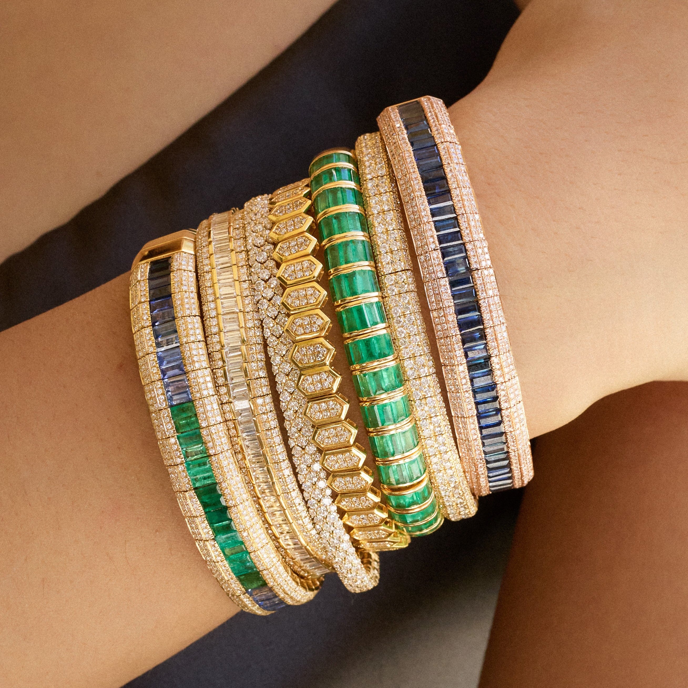 Close-up of a wrist adorned with a stack of luxurious diamond, emerald, and sapphire bracelets in yellow gold and rose gold settings